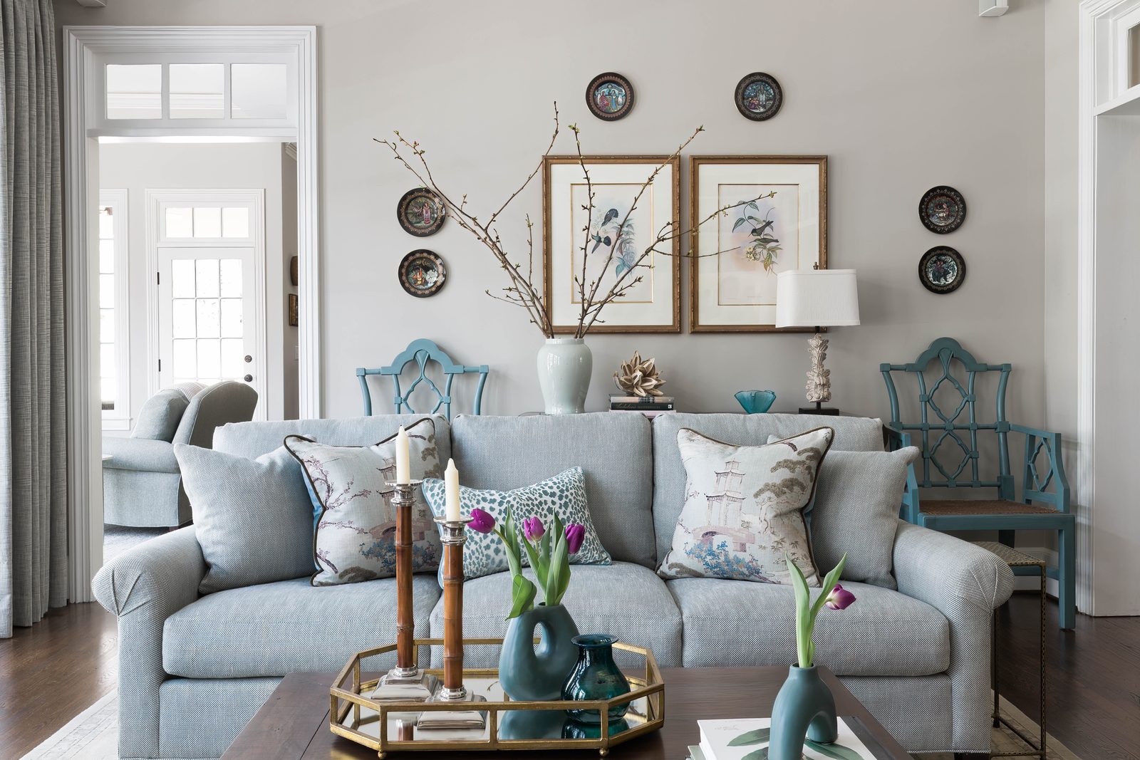 Wide editorial interior photograph of a soft gray living room with patterned blue armchairs, a watercolor diptych above the sofa, fresh tulips on a brass tray — a frame built around asymmetric composition and considered focal layering.