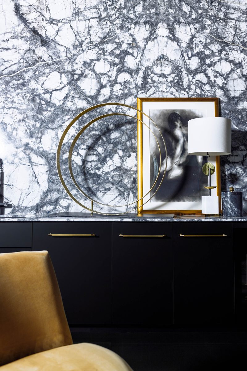 Vertical editorial detail of a console vignette — black marble waterfall counter, brass-ringed sculptural object, a framed black-and-white figure study, and a brass-and-marble lamp — demonstrating focal-point restraint and asymmetric framing.
