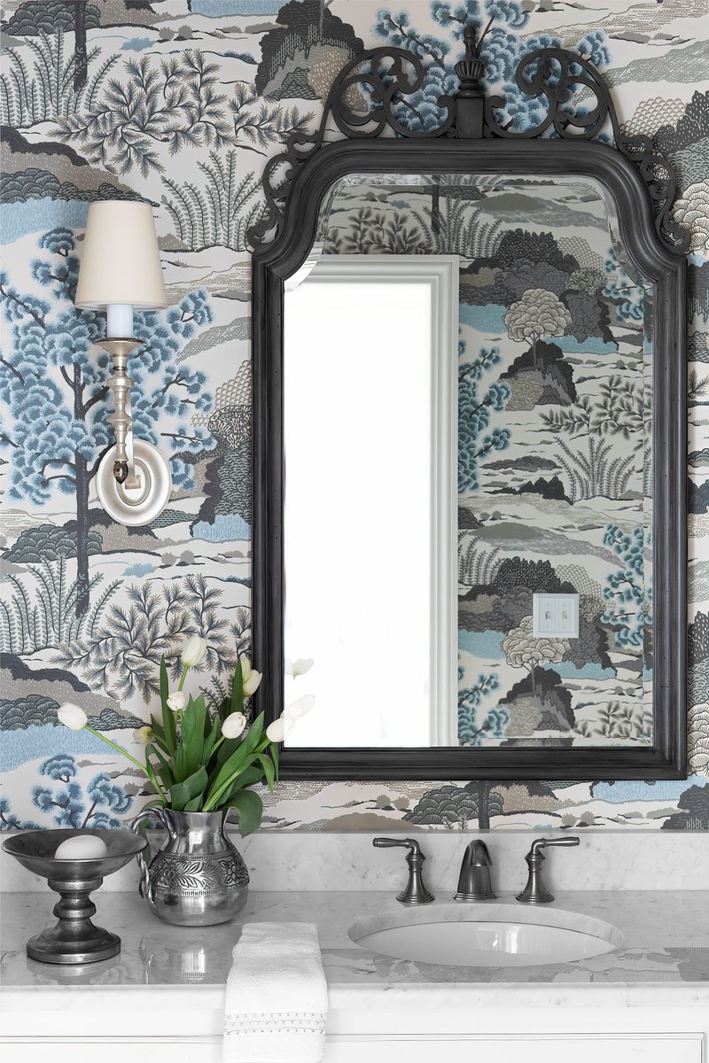 Vertical editorial detail of a powder room — a dark-framed ornate mirror reflecting a window, an antique-style sconce, white tulips in a pewter pitcher on a marble counter, against a blue-and-cream scenic wallpaper — exactly the vertical-format frame shelter magazines use for lead spreads.