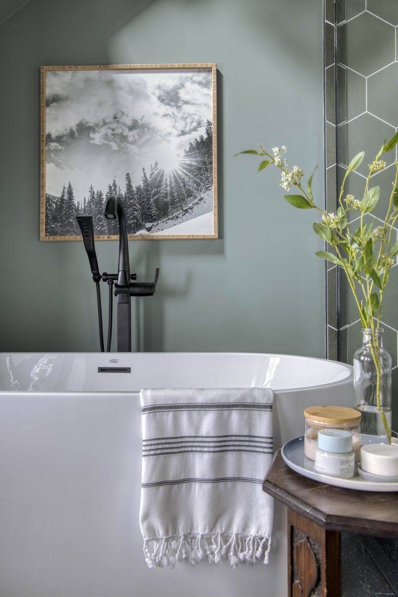 Vertical editorial detail of a freestanding tub with a fringed striped hand towel draped over the rim, fresh florals in a glass vase, and a framed landscape print — the textile and surface styling decisions that show up immediately through the lens.
