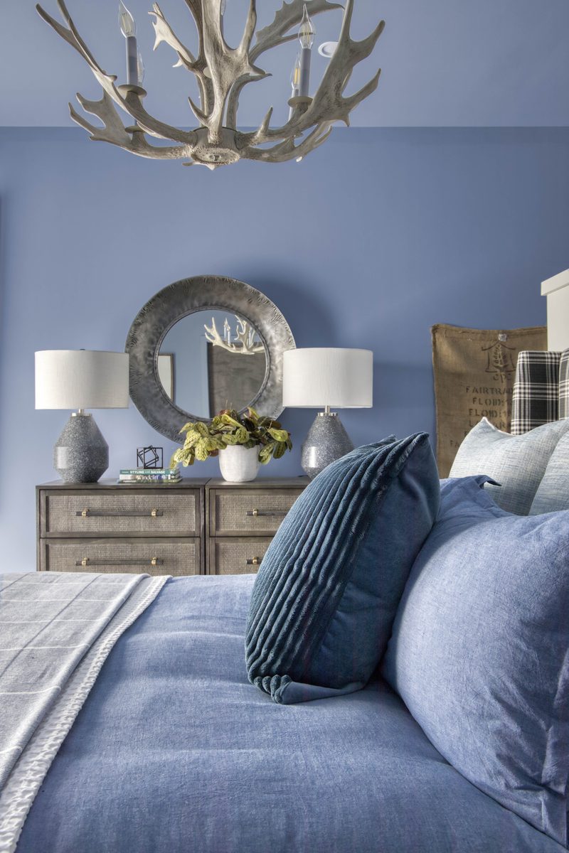 Vertical editorial interior photograph of a periwinkle bedroom with an antler chandelier, twin lamps on a textured dresser, a round mirror, and layered blue and tartan bedding — a textile-heavy frame where every styling decision is visible to the camera.