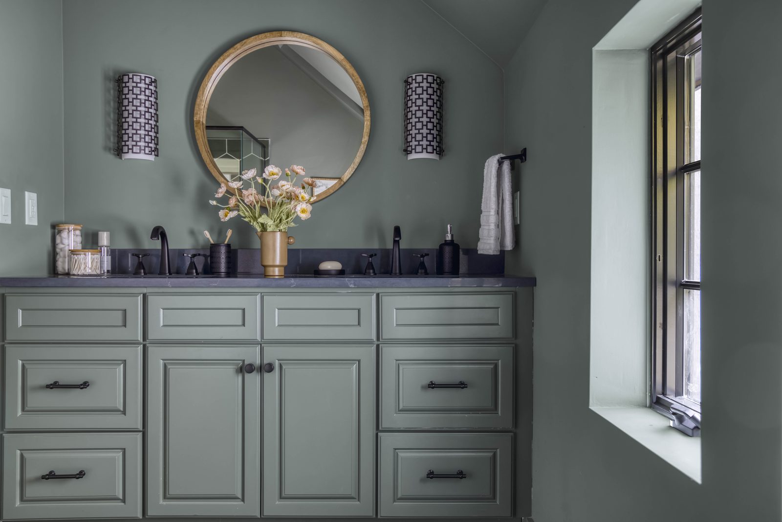 Editorial interior photograph of a sage-green powder room vanity with a round mirror, twin patterned sconces, a single arrangement of cream daisies, and dark stone counter — restrained styling with one focal point, the visual equivalent of 'when in doubt, take it away'.