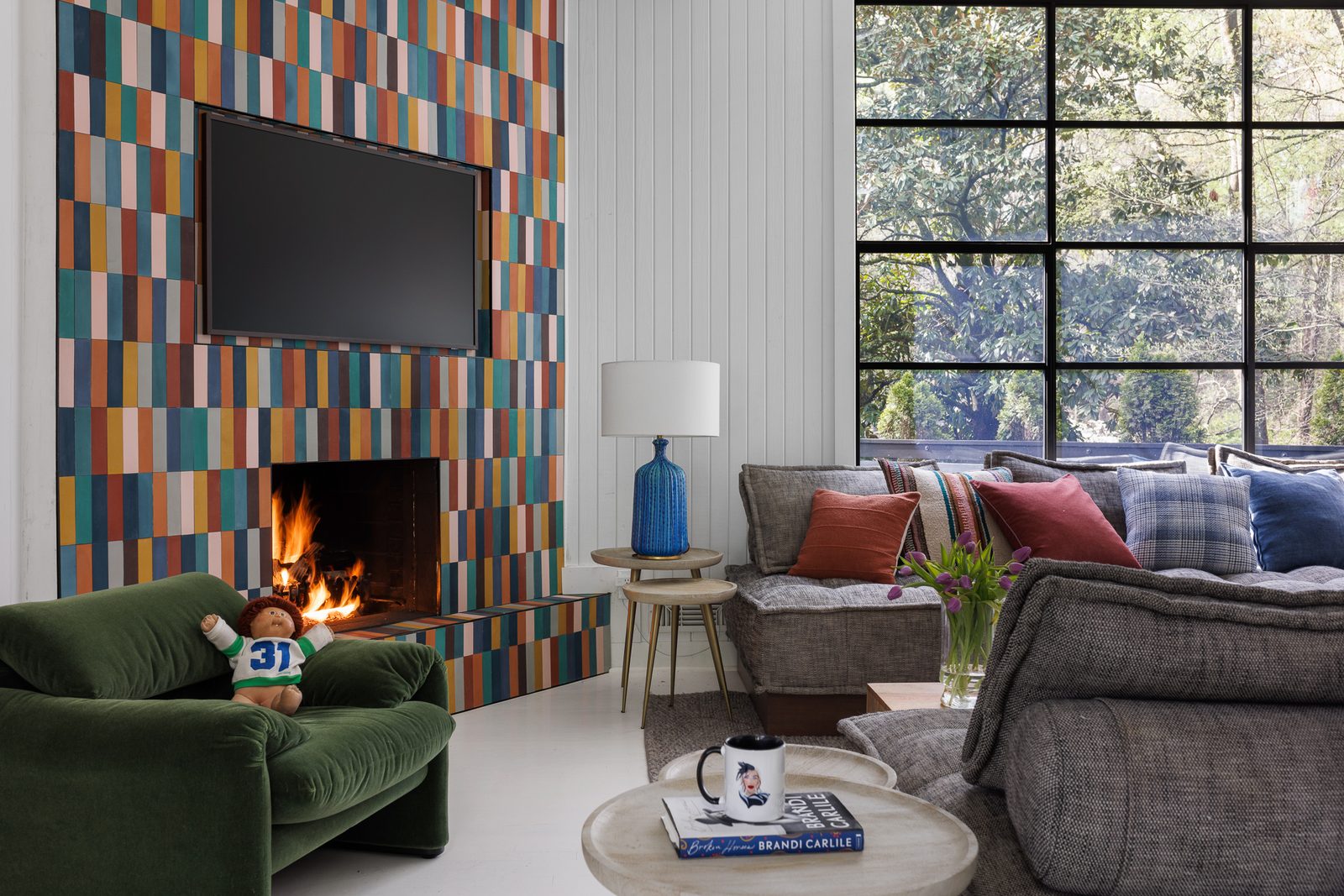 Publication-ready editorial interior photography of a colorful living room with multicolor tile fireplace, layered textiles, and floor-to-ceiling windows