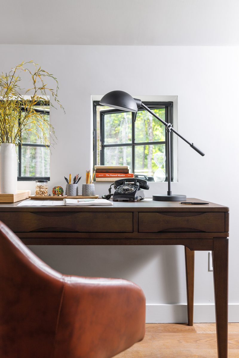 Vertical editorial photograph of a styled home office — walnut desk with a vintage black telephone, brass desk lamp, books propped against a window, fresh greenery in a white vase, and a leather chair pulled in — a frame that reads as prepared down to the smallest surface detail.
