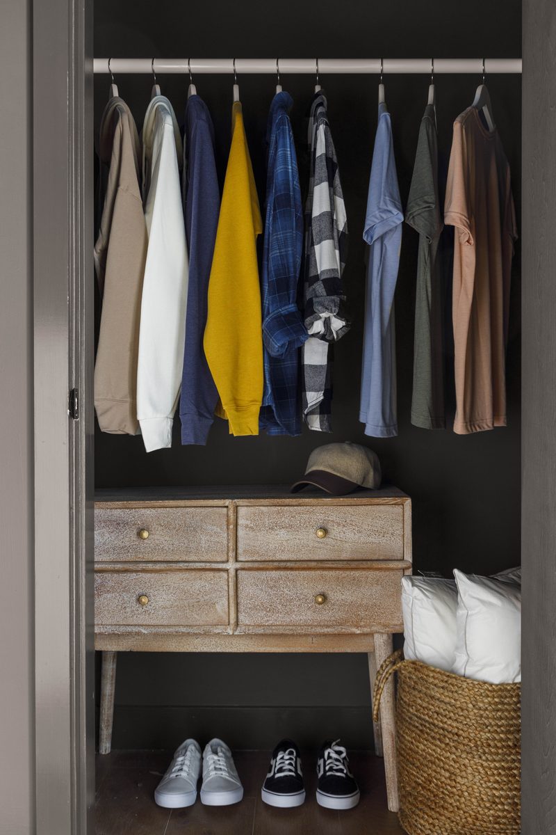 Vertical editorial photograph of an open custom closet — a row of intentionally arranged garments on matching hangers, a four-drawer console with brass pulls, a folded blanket and woven basket below, and two pairs of sneakers on the floor — visual shorthand for the kind of organized infrastructure a well-built shoot kit represents.