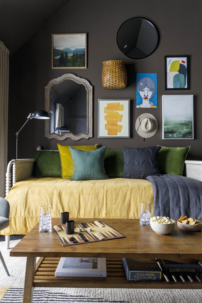 Vertical editorial photograph of a sitting room with charcoal walls, a gallery of mixed art and a woven basket above a yellow-upholstered daybed, layered with green, mustard, and slate pillows and a charcoal throw — exactly the kind of textile-heavy frame where steamed fabric makes or breaks the image.